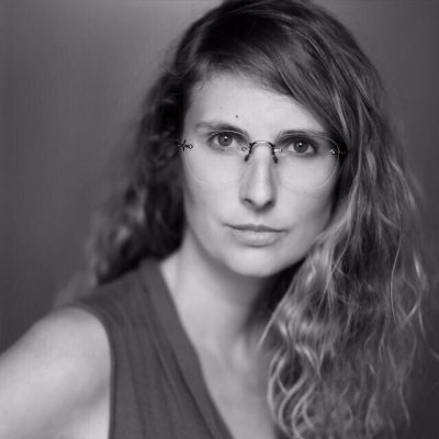 Porträt einer Frau mit Locken und Brille, ernster Blick, einfarbiger Hintergrund.