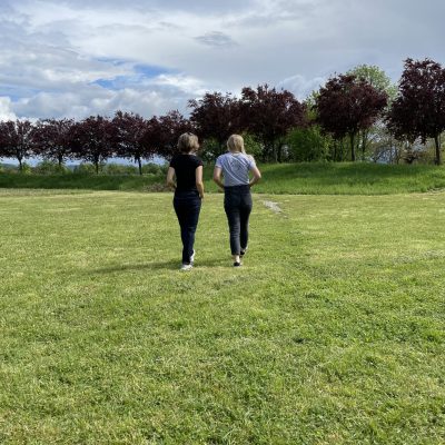 Zwei Personen gehen auf einer Wiese, umgeben von Bäumen unter bewölktem Himmel.