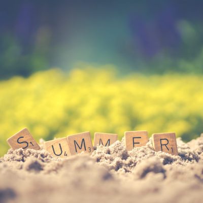 Scrabble-Buchstaben bilden das Wort 'SOMMER' auf Sand, mit einem Feld gelber Blumen im Hintergrund.